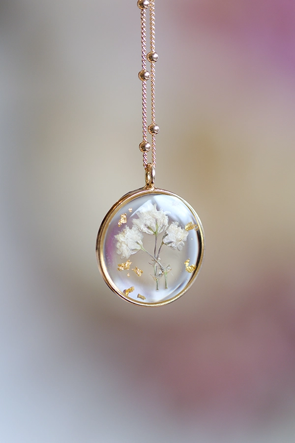 Podstavek ovalne oblike iz prozorne epoksi smole, v katero so ujeti delčki posušenih cvetov v modri in rožnati barvi, ter lističi 24-karatnega zlata.